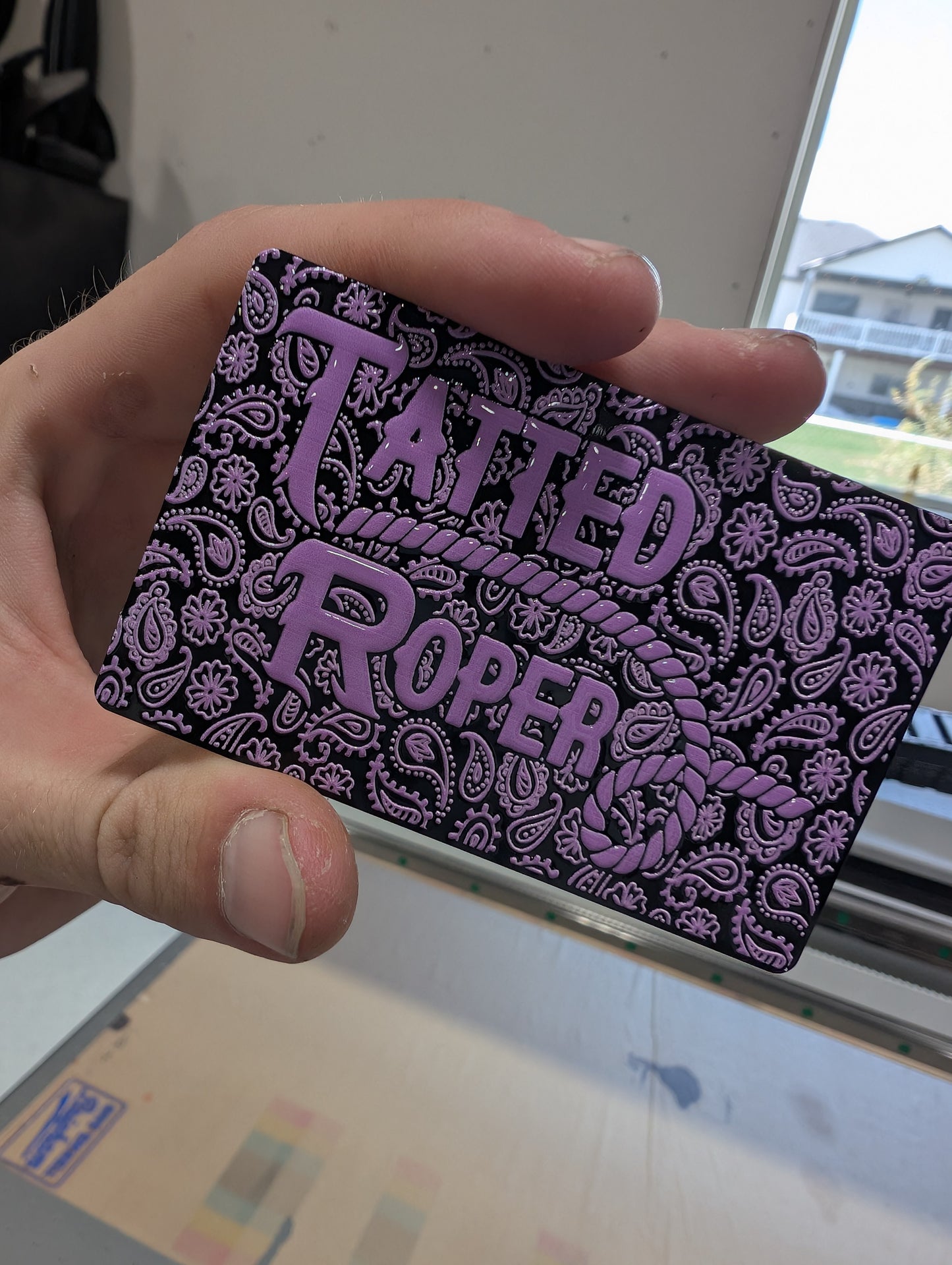 Hand holding a card with 'Tatted Roper' design in purple on a black background